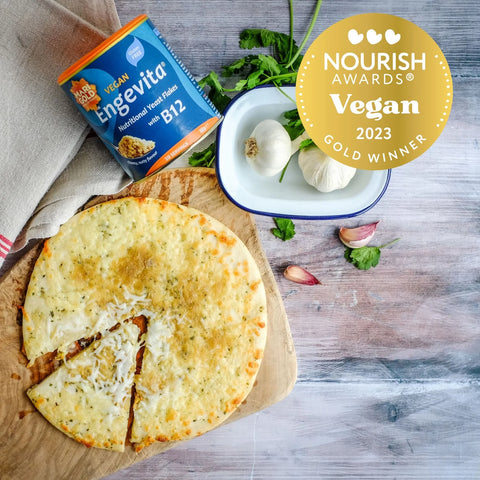 A sliced round vegan bread on a wooden board with a can of Engevita plant-based bread flakes, a white dish with garlic, parsley, and cloves, and a gold Nourish Awards Vegan 2023 Winner badge on a rustic gray wood surface.