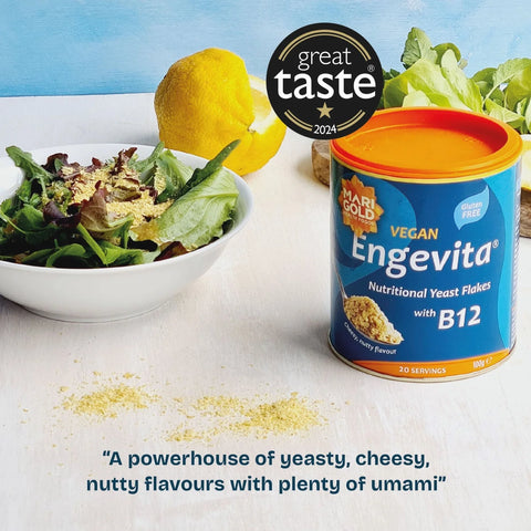 A can of Hari Gold Enevita Vegan Nutritional Yeast Flakes with B12 stands on a white surface beside a bowl of mixed greens and a lemon, with a black 'great taste 2024' badge above and a quote at the bottom.