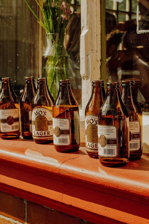Several brown glass beer bottles with white labels reading 'MO'S LAGER' lined up on a red-orange windowsill in warm natural light.