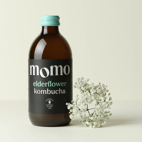 An amber glass bottle of Momo Elderflower Kombucha with a black label and a light blue cap, styled next to a small cluster of white baby's breath flowers on a light beige background.