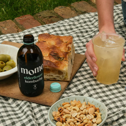 A bottle of momo elderflower kombucha sits on a picnic blanket beside a slice of rustic bread, bowls of olives and nuts, and a hand holding a glass of iced kombucha outdoors.