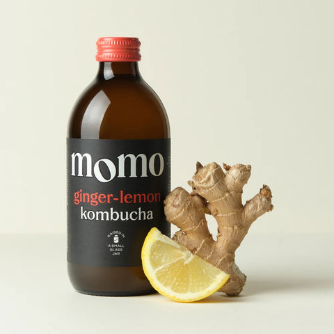 An amber glass bottle of Momo Ginger-Lemon Kombucha with a red cap and black label, styled with a slice of lemon and a piece of ginger root on a light beige background.