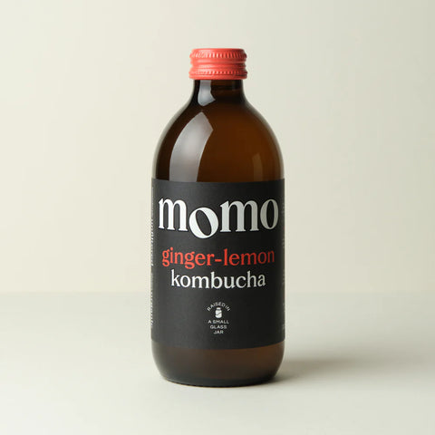 A bottle of Momo Ginger-Lemon Kombucha in an amber glass bottle with a black label and a red cap, set against a plain light beige background.
