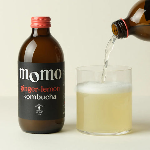 A bottle of Momo Ginger-Lemon Kombucha stands next to a clear glass being filled with the pale yellow kombucha liquid from an amber bottle, on a light beige background.