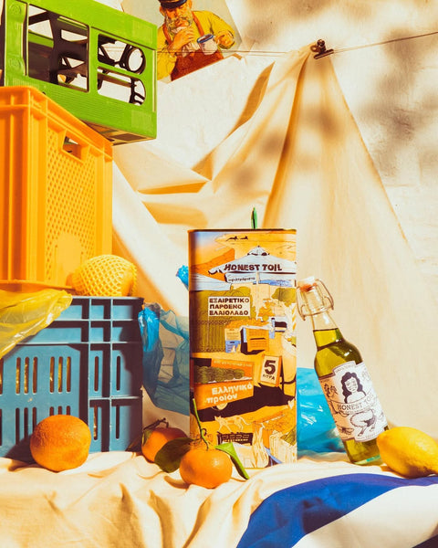 Honest toil 5l olive oil tin with 1l bottle leaning against with citrus fruit spread around them