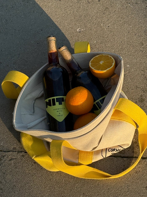 A beige canvas tote bag with bright yellow handles sits on concrete outdoors, filled with two dark wine bottles, two whole oranges, and a halved orange. The bag has a visible partial brand print reading 'CORM.