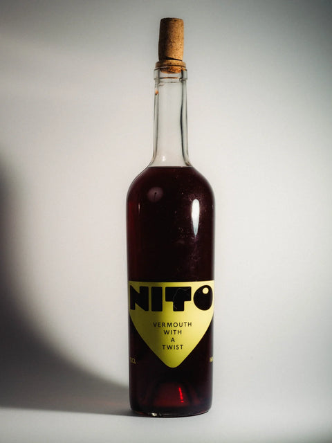 A clear glass bottle of Nito Vermouth with a Twist, filled with dark red liquid and sealed with a cork stopper, set against a neutral gray background.