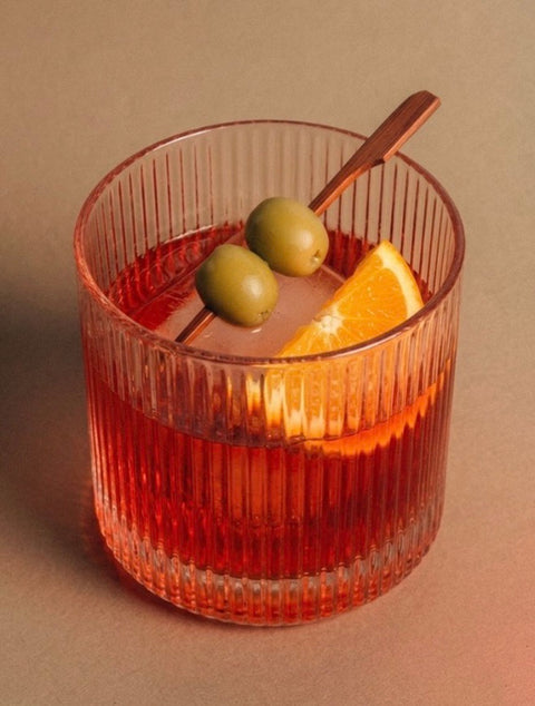 A clear ribbed tumbler glass containing an orange-red drink, garnished with two green olives on a metal skewer and an orange slice, set against a warm beige background.