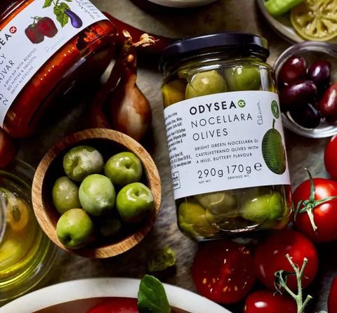 A glass jar of Odyssea Nocellara Olives prominently displayed on a rustic table, surrounded by fresh green olives in a wooden bowl, red tomatoes, olive oil, and another jar of Odyssea pasta sauce.