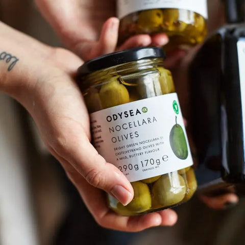 A hand holds a glass jar of Odyssey Nocellara Olives, with another jar in the background and a dark bottle partially visible on the right.