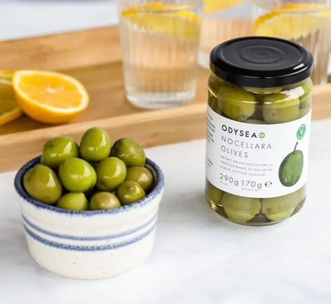 A jar of Odyssey Nocellara Olives next to a small bowl of fresh green olives on a white marble surface, with a wooden board, sliced oranges, and glasses of water in the background.