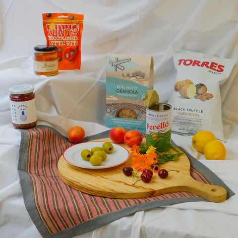 Assorted food items including a plate of grapes, a jar of jam, a bag of granola, and a can of Perello wine on a wooden board with a white background.