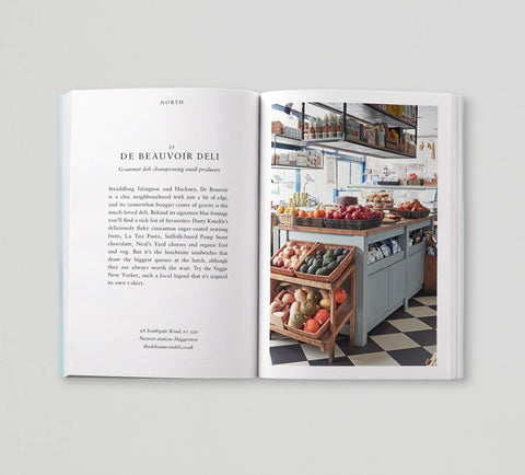 An open printed magazine or book spread photographed against a light gray background. The left page contains a recipe or food article titled 'DE BEAUVOIR DELI' with text only, and the right page shows a color photograph of a deli counter with fruit, shelves, and a checkered floor.