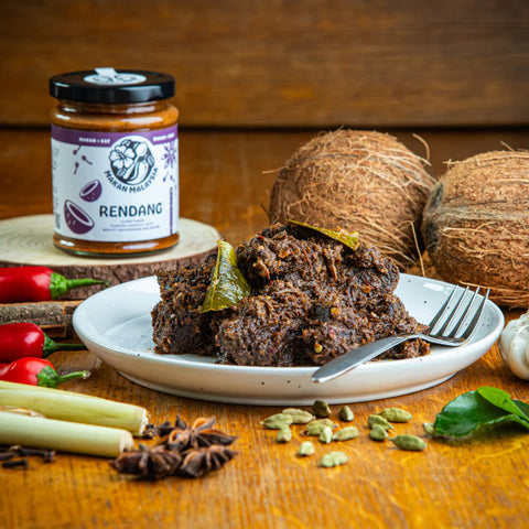 Plated dish of Rendang with coconuts, peppers, and a jar labeled 'Rendang' on a wooden surface.
