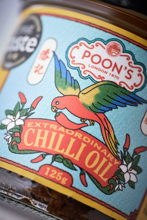 Close-up of a glass jar of Poon's London Extraordinary Chilli Oil, showing the colorful vintage-style label with a red, yellow, and blue parrot on a light blue background.