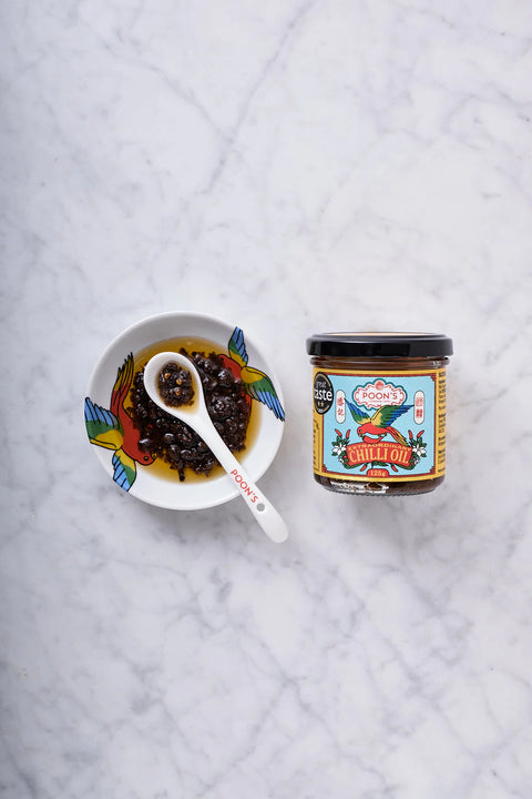 A glass jar of Siam Food Company Sweet Chili Sauce next to a small white bowl containing cooked chili paste with a white spoon, set on a white marble surface.
