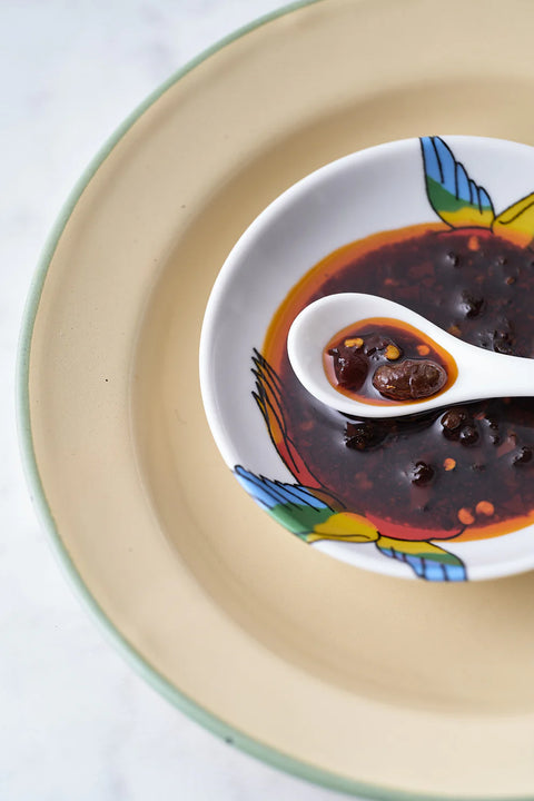 A close-up of a white ceramic bowl with colorful painted bird motifs, filled with a dark reddish-brown sauce or chili oil, with a white spoon resting in it. The bowl sits on a beige plate against a light marble-like background.