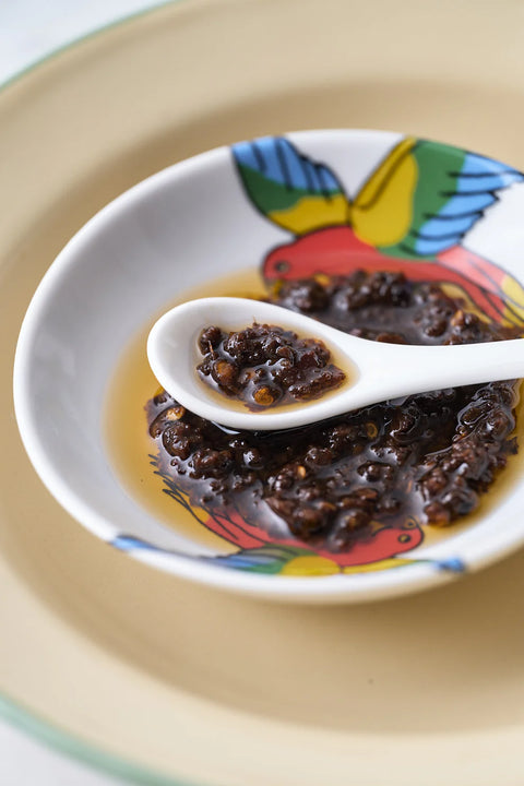 A close-up of a white ceramic bowl with a colorful bird pattern, filled with a dark, chunky sauce or relish on a light beige plate, with a white spoon holding some of the sauce.