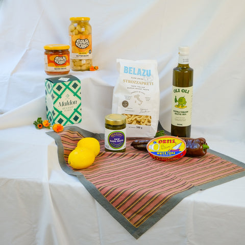 Assorted food products including a bottle of olive oil, a jar of pickles, and a bag of pasta on a white tablecloth.