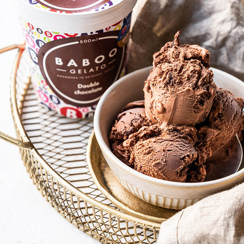 A bowl of chocolate gelato ice cream next to a tub of BABOO Gelato Double Chocolate on a gold tray.