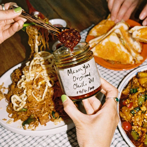 Person holding a jar of 'Mama Yu's Original Chilli Oil' with food in the background