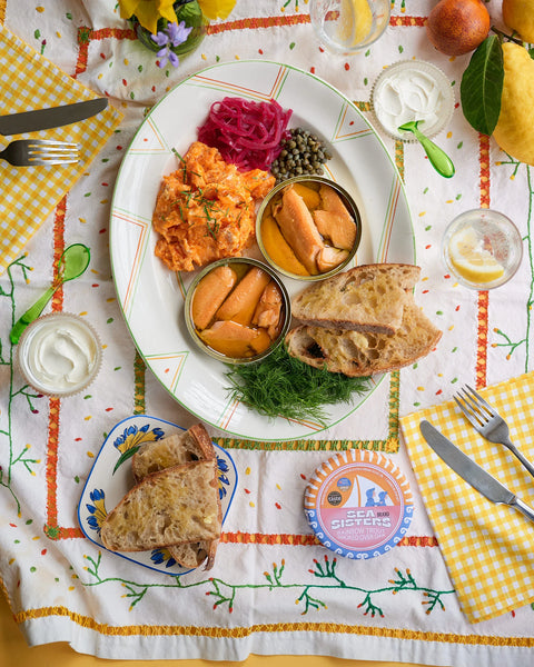 A top-down view of a breakfast spread on a white tablecloth with yellow gingham napkins. The main plate holds smoked salmon, pickled red cabbage, capers, and dill, alongside a small plate with a slice of toast.
