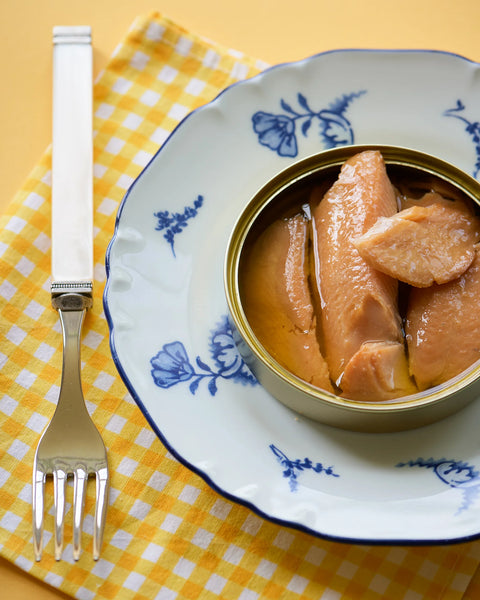 An open can of smoked rainbow trout in oil, served on a blue and white patterned ceramic plate with a yellow and white checkered napkin and a silver fork on a yellow table.