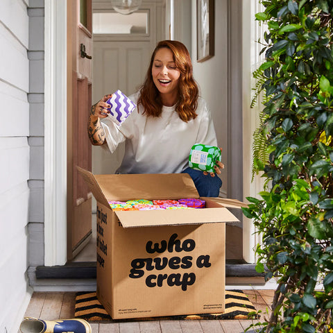 A smiling woman stands in a doorway holding a large open cardboard box filled with colorful packaged items. She holds a purple-and-white patterned item in one hand and a green item in the other.