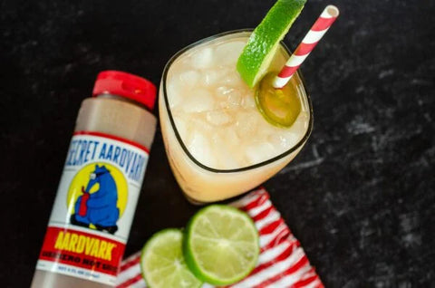 Cocktail with lime and straw next to a bottle labeled 'Secret Aardvark' on a dark background