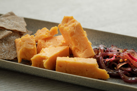 Slices of orange red leicester cheese arranged on a dark rectangular serving platter alongside crackers and pickled red onions on a light wood surface.