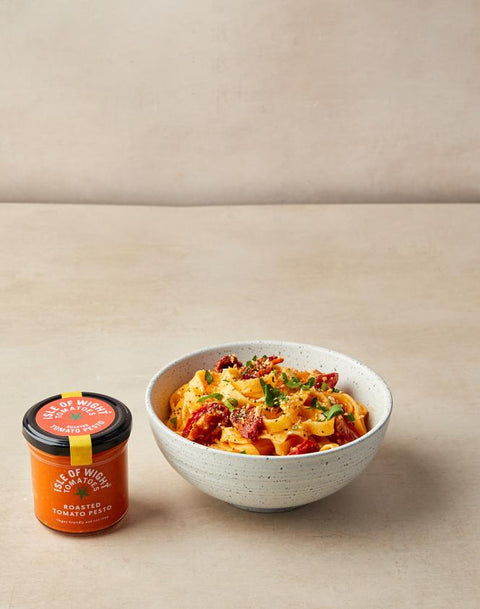 A bowl of creamy tomato pasta with sunburst tomatoes and green herbs, served next to a small jar of 'Tis of Witch' Roasted Tomato Sauce on a neutral beige surface.