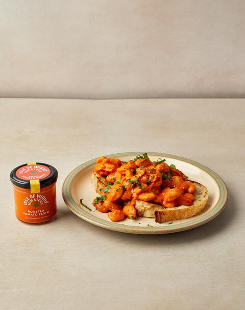 A small jar of orange-red spread sits beside a plate of bread topped with a chunky orange-red sauce and chopped green herbs, photographed on a light beige surface against a matching neutral background.