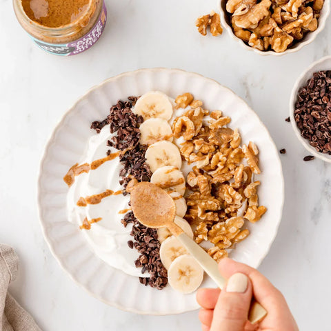 A top-down view of a white plate of yogurt topped with sliced bananas, walnuts, cacao nibs, and drizzles of peanut butter sauce. A hand is holding a spoon with peanut butter, and bowls of ingredients are arranged around the plate on a light marble surface.