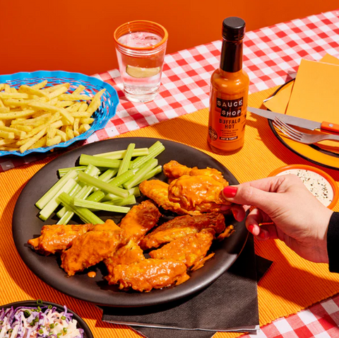 plate of wings and celery next to bowl of chips and cup of water on red checkered surface. Bottle of sauce shop buffalo hot.