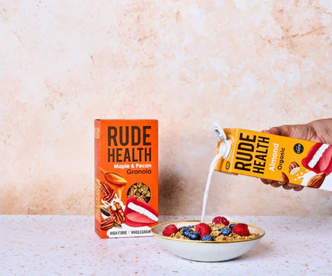 A hand holds a yellow Rude Health Organic Granola carton while pouring milk into a bowl of granola with berries, next to a larger orange Rude Health granola box on a light speckled surface against a textured beige wall.