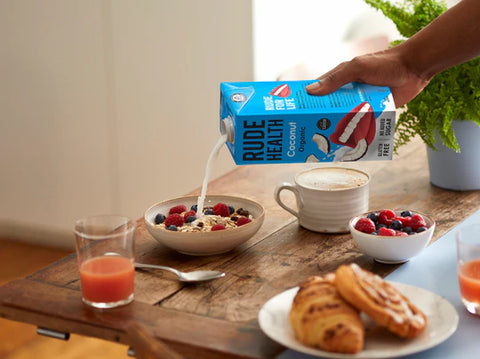 A hand holds a blue carton of Rude Health Coconut Almond Milk while pouring it into a bowl of cereal on a wooden table, with a cup of coffee, fruit, and pastries nearby.