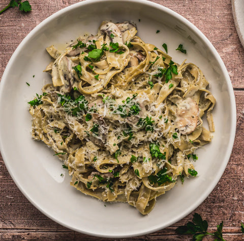 A top-down view of a creamy pasta dish served in a white bowl on a rustic wooden table, garnished with fresh parsley and grated cheese.