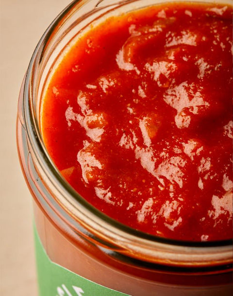 A close-up shot of a glass jar filled with chunky red tomato sauce, showing the glossy, textured surface of the sauce inside a clear glass container.