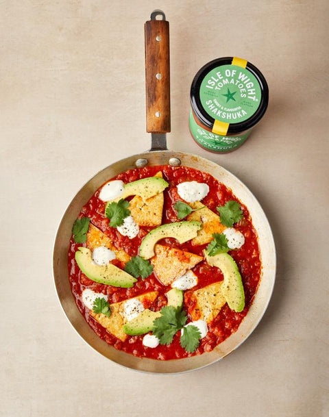 A top-down view of a cast iron skillet filled with a red tomato-based stew or shakshuka, topped with avocado slices, white sauce, and cilantro, next to a jar of Pick of Midnight Habanero Chiles on a beige background.