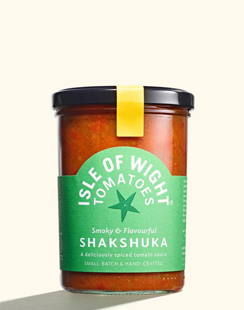 A glass jar of Isle of Wight Tomatoes Shakshuka, featuring a green label and a black lid with a yellow seal, set against a solid cream background.