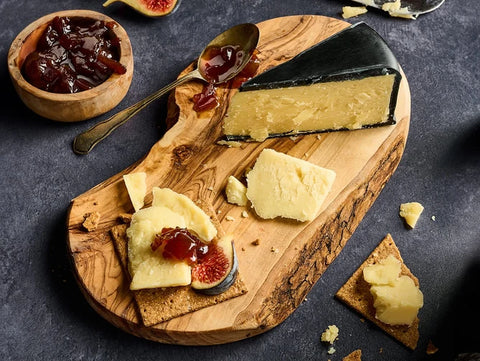 A styled food scene showing a wedge of pale yellow cheese with a dark rind on a wooden serving board, alongside figs, crackers, a small bowl of dark red jam, and a spoon with jam on a dark textured surface.
