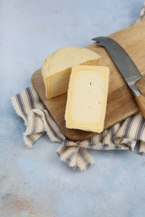 Two wedges of pale yellow cheese on a wooden cutting board with a knife, set on a striped cloth over a light blue surface.