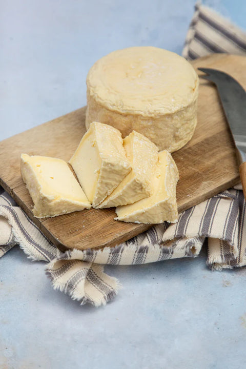 A round wheel of soft yellow cheese with several wedges cut in front, displayed on a wooden cutting board with a striped cloth and a knife on a light blue surface.