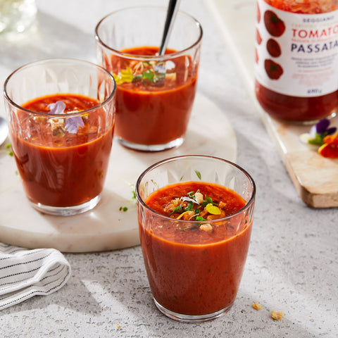 Three glasses of red tomato soup garnished with herbs and nuts are arranged on a light stone surface, with a bottle of Reggiani Passata tomatoes in the background.