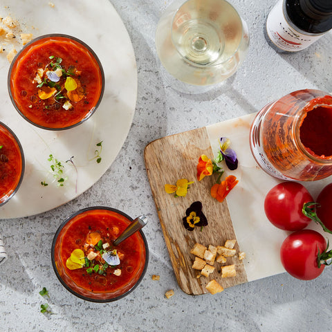 Overhead flat-lay of a tomato-based soup or gazpacho dish, with two clear glasses filled with red soup topped with herbs and edible flowers, a jar of red sauce or tomato product, whole tomatoes, a glass of white wine, and a cutting board on a light stone surface.