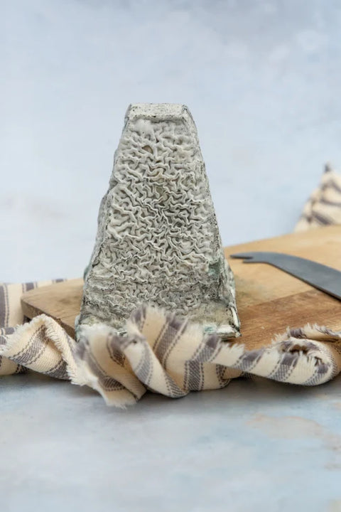 A wedge of hard blue cheese standing upright on a wooden cutting board, with a striped cloth and a knife nearby, against a soft light gray background.