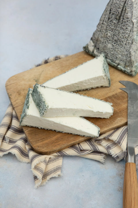 Three wedges of soft white cheese with blue-gray rind resting on a wooden cutting board, with a whole wedge and a cheese knife nearby on a light blue surface.
