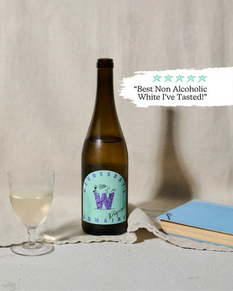 A dark glass bottle of Wednesday Domain Non-Alcoholic White wine with a light blue and purple label, styled on a textured beige surface next to a glass of white wine, a blue notebook, and a star rating graphic with the quote 'Best Non Alcoholic White I've Tasted!'.