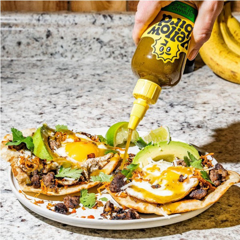 A hand holds a bottle of yellowbird Hot Sauce, pouring the yellow sauce over two loaded tortilla chips on a white plate, set on a granite countertop.