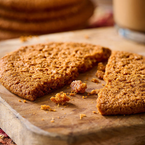 A close-up of a golden-brown oatmeal cookie broken in half on a wooden cutting board, with crumbs scattered around and a blurred mug in the background.